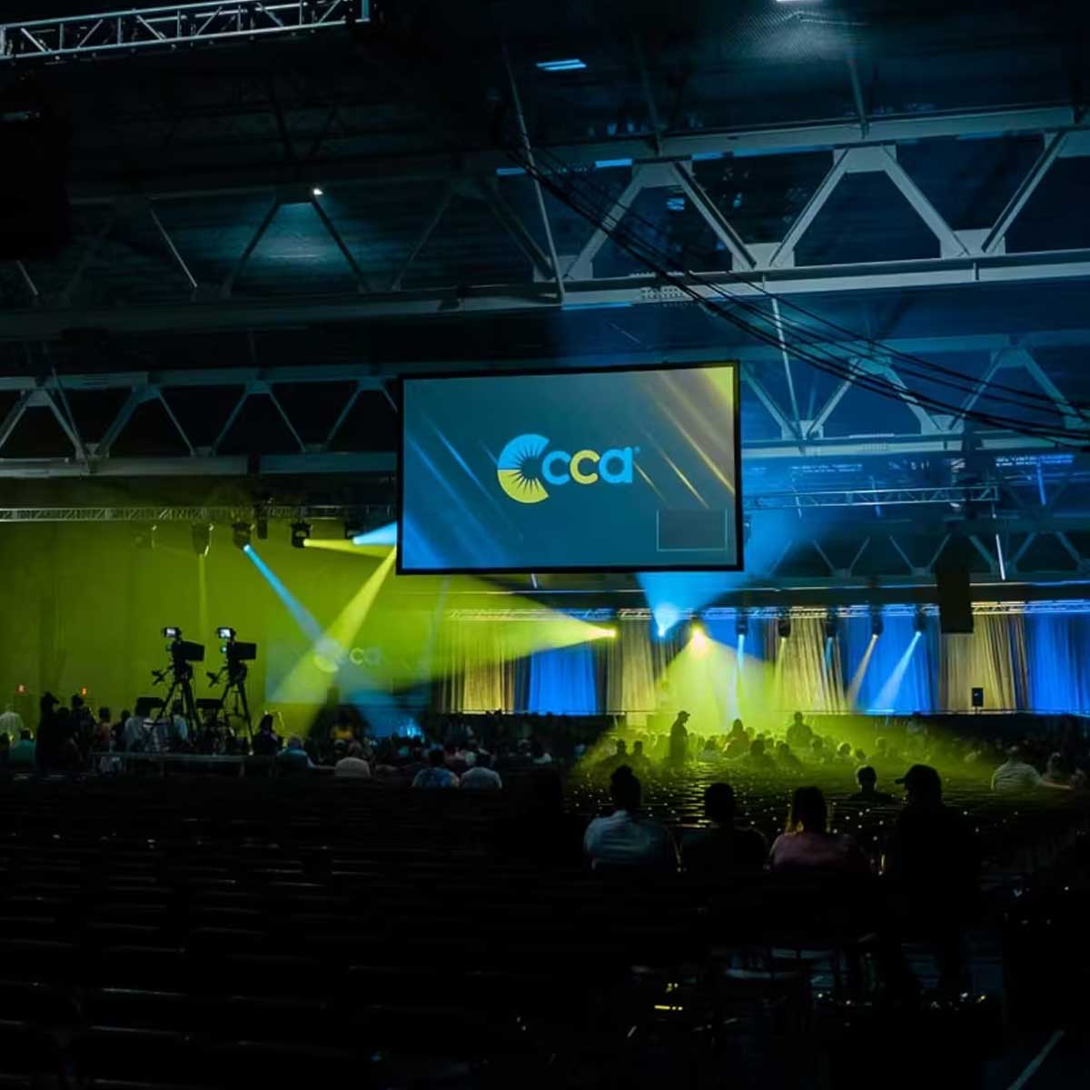 Large indoor event space with stage, blue and yellow lighting, and CCA logo on screen