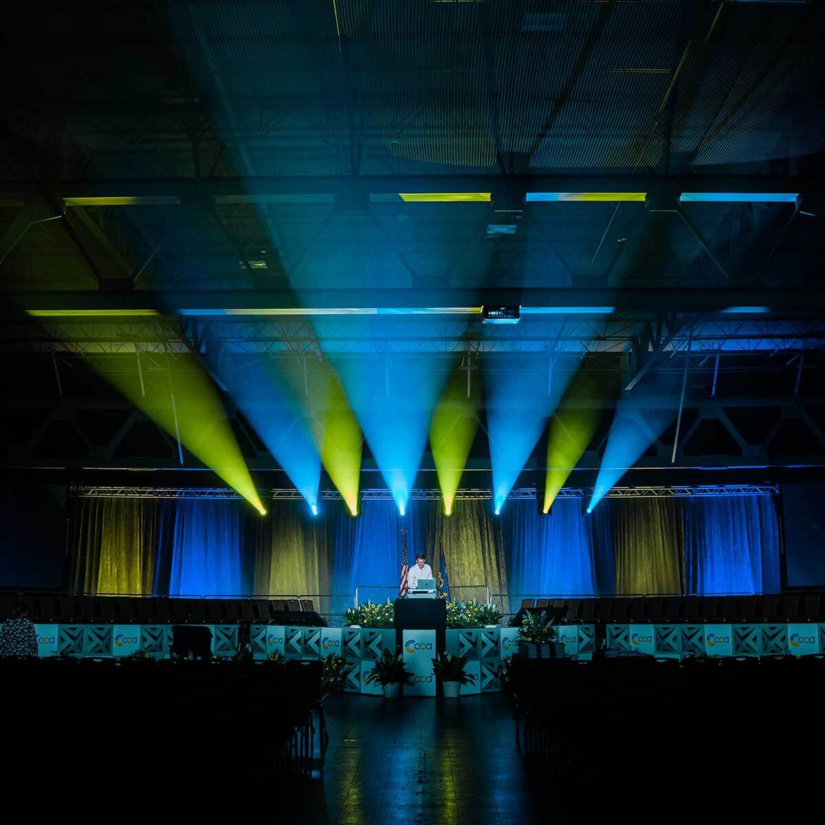 A person stands at a podium on a stage, illuminated by blue and yellow spotlights
